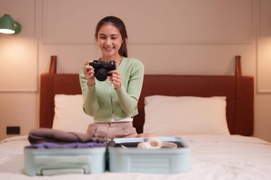 Bir otel odasında bavulunu toplayan gülümseyen genç bir Asyalı kadın. Heyecan verici tatil macerası için elinde bir kamera tutuyor..