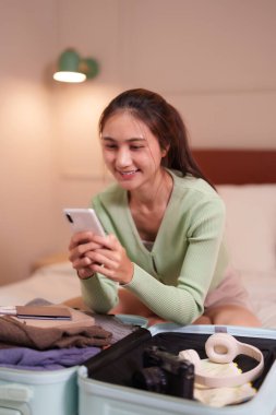Gülümseyen Asyalı kadın, yaklaşan sıcak yatak odası seyahati için bavullarını hazırlarken mobil cihaz kullanıyor..