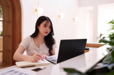 Genç Asyalı yetişkin bir kadın dizüstü bilgisayara odaklanmış ve serbest meslek eğitimi için ideal, modern bir kafede notlar yazıyor.