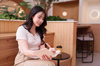 Genç Asyalı kadın, çağdaş bir kafede, bir deftere kahve eşliğinde yaratıcı bir çalışma seansının tadını çıkarıyor.