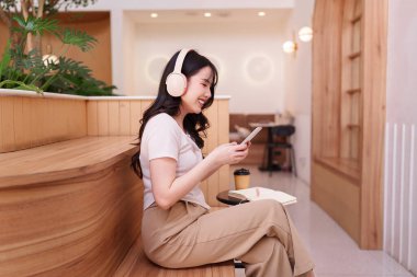 Asyalı kadın akıllı telefon, kablosuz kulaklık, rahat kafe seti kullanıyor. Ses içerikli dijital iletişimden hoşlanıyor. Rastgele uzaktan kumanda deneyimi.