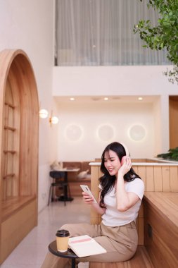 Genç Asyalı kadın canlı sesli kafe ortamını seviyor. Gülümseyen kadın akıllı telefonla iletişim kurar. Rahatlamak ya da çalışmak için rahat çalışma alanı.