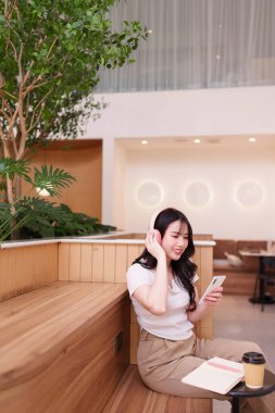 Asyalı yetişkin kadın akıllı telefon kafede kulaklıkla ses dinliyor. Modern sakin ortam mükemmel dijital yaşam tarzı rahatlaması.