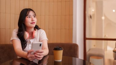 Asyalı genç yetişkin, modaya uygun bir kafede huzurlu bir anın tadını çıkarıyor. Mobil aygıtı tutuyor. Kulaklıklar sıcak içeceğin yanında..