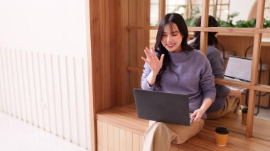 Genç Asyalı kadın, modern kafeterya dostu dijital göçebe yaşam tarzında dizüstü bilgisayarla video görüşmesinde gülümsüyor.