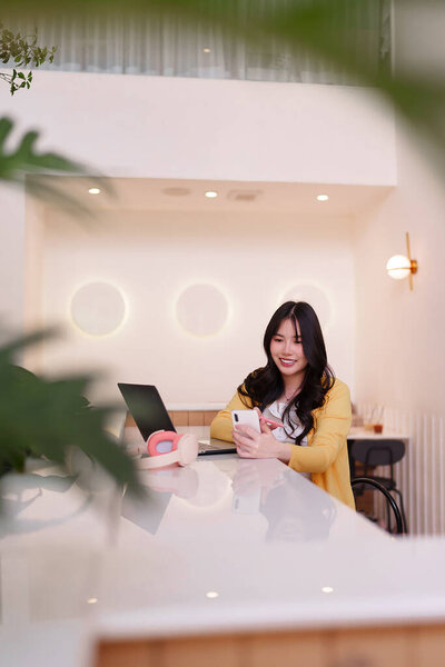 Взрослая азиатка удаленно работает в ярком кафе. Она улыбается, используя смартфон и ноутбук для онлайн-бизнеса..