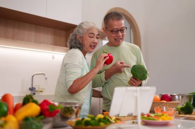 Neşeli Asyalı yaşlı yetişkinler besleyici yemekleri ev mutfağında hazırlıyorlar. Canlı ve sağlıklı yaşam için tabletteki dijital tarifleri takip ediyorlar..