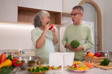 Neşeli yaşlı Asyalı erkek kadın sağlıklı beslenme tarzını geliştirmek için açık modern mutfakta internetten taze sebzeler hazırlıyor..