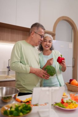 Neşeli Asyalı yaşlı yetişkinler, taze ürünleri olan parlak çağdaş bir mutfakta birlikte yemek yapmayı severler. Besleyici yemekler hazırlarlar.