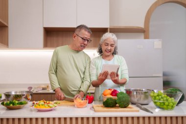 Mutlu Asyalı yaşlı çift, ev mutfağında besleyici yemekler hazırlıyor. Erkek dilimleri, internet üzerinden kadın tarifi üretiyor. Sağlıklı yaşam tarzı kavramı.