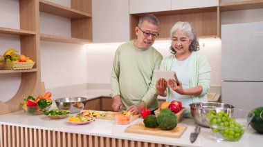 Neşeli Asyalı yaşlı çift taze sebze pişiriyor. Parlak modern mutfakta. Sağlıklı yaşam için dijital tarif tabletine göz atıyorlar.