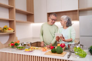 Asyalı yaşlı çift, modern mutfakta sağlıklı yemekler pişirmek için ilham verici dijital tablet kullanıyor. Taze sebzeler hazırlanmayı bekliyor.
