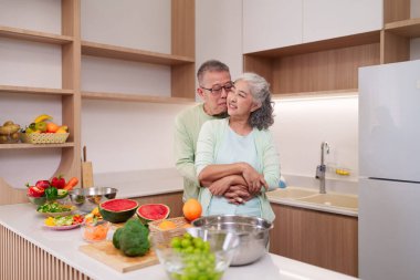 Sevecen Asyalı yaşlı çift, sevgi dolu bir anı paylaşıyor. Parlak bir ev mutfağında besleyici yemekler hazırlıyor. Sağlıklı yaşam tercihlerini birlikte destekliyorlar.