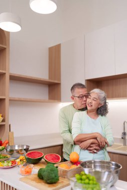 Mutlu Asyalı yaşlı yetişkinler kucaklaşır birlikte taze ve sağlıklı yiyecekler hazırlarlar. Modern mutfak sahnesinde sağlıklı yaşam ve sevgi dolu bir ilişki sergileniyor.