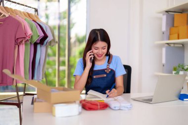 Young adult Asian entrepreneur communicates customers processes clothing orders home office. E-commerce business management daily tasks.