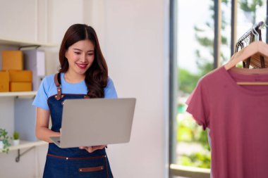 Happy Asian woman entrepreneur operates online apparel business. Manages orders marketing customer support on laptop from home office