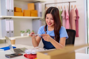 Young Asian woman business owner photograph merchandise for online store. Fulfill orders pack boxes at home office for customers.