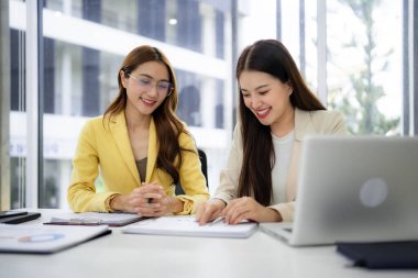 Two smiling professional women engage in productive office discussion reviewing important paperwork. They work together analyzing data for strategic decision making.