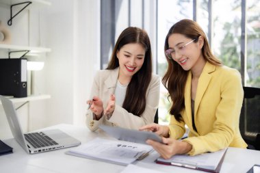 Asian female colleagues analyze documents. They discuss important corporate project plans. Positive work atmosphere encourages innovation.