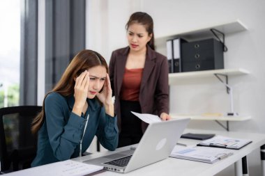 Two Asian adult colleagues confront workplace stress. One woman appears overwhelmed by a problem while her manager discusses documents. Professional office setting illustrates work pressure.