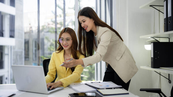 Two Asian female professionals engage in productive discussion reviewing project data on a laptop in a bright modern office workspace