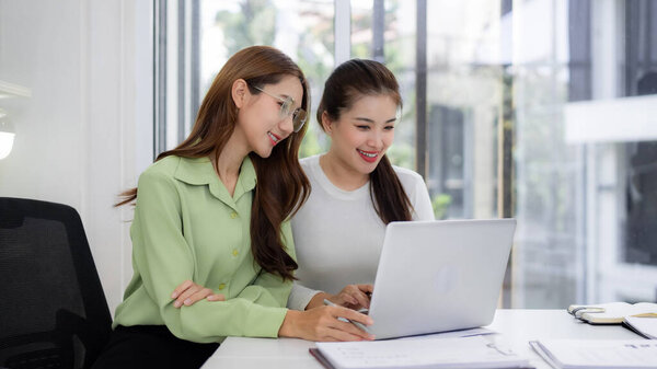 Two Asian adult women share ideas analyzing data on a laptop in a bright contemporary office setting demonstrating teamwork and productive discussion