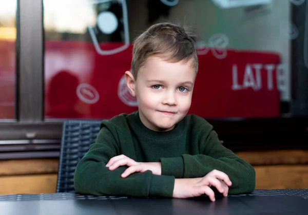 The boy sits at the table and looks straight ahead. The light-haired boy is 6 years old. He is wearing a green sweater and smiling. The photo is blurred. High quality photo