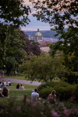 Gençler piknik yapıyor ve 22 Temmuz 2024 'te Çek Cumhuriyeti' nin başkenti Prag 'ın Vinohrady ilçesindeki Riegrovy Sady parkında Prag şehir manzarasının tadını çıkarıyor.