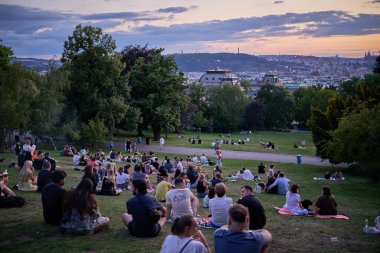 Gençler piknik yapıyor ve 22 Temmuz 2024 'te Çek Cumhuriyeti' nin başkenti Prag 'ın Vinohrady ilçesindeki Riegrovy Sady parkında Prag şehir manzarasının tadını çıkarıyor.