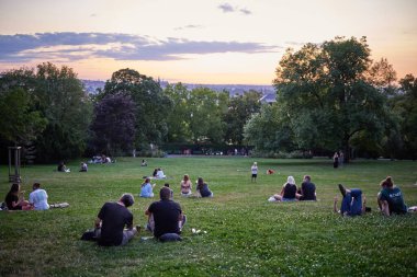 Gençler piknik yapıyor ve 22 Temmuz 2024 'te Çek Cumhuriyeti' nin başkenti Prag 'ın Vinohrady ilçesindeki Riegrovy Sady parkında Prag şehir manzarasının tadını çıkarıyor.