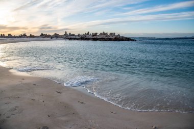 Sahil manzaralı sakin deniz, kumlu sahil ve gün batımında canlı bir gökyüzü altında kayalık oluşumlar.