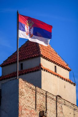 Açık mavi gökyüzünün altındaki tarihi tuğla kulede dalgalanan Sırp bayrağı