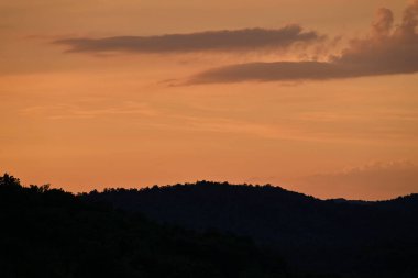 A serene sunset with orange and purple clouds over a silhouetted forested hill.