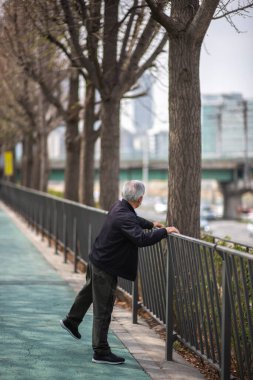 Yaşlı adam 25 Mart 2023 'te Güney Kore' nin Seul kentsel yolu boyunca bir korkuluğa yaslanarak egzersiz yapıyor.
