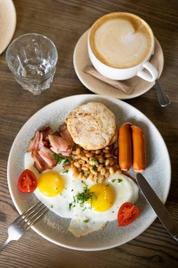 İngiliz kahvaltısı. Sosisli sahanda yumurta, fasulye, pastırma ve domatesli beyaz tabak ve bir fincan kahve.