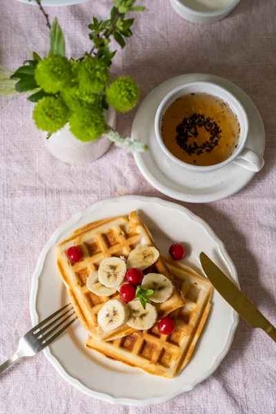 ホワイトプレートにバナナとチェリーのベルギーワッフル 紅茶と花のカップ 甘い朝食 — ストック写真