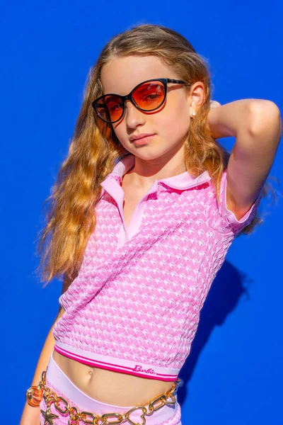 Attractive model Barbie style girl wearing sunglasses, pink tee-shirt and shorts with long hair on blue backdrop, vintage fashion concept. Hight quality photo.
