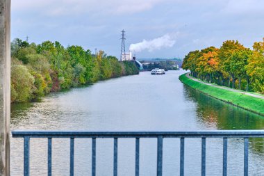 Manzaranın geniş bir kanalı var. Köprünün üstünden temiz su akıyor. Uzakta tırabzan var. Kanalda büyük bir gemi.