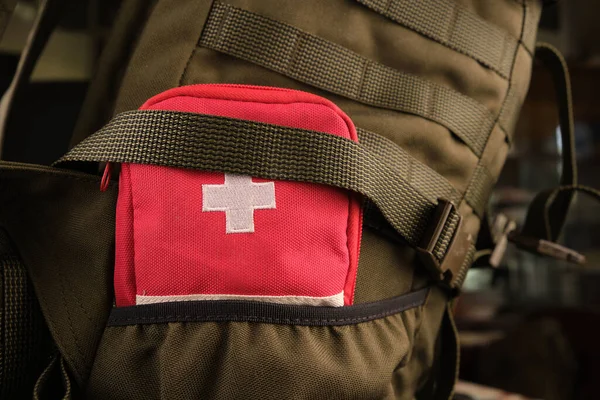 A red first aid kit with a white cross in the pocket of a tactical military olive backpack with a Molle system