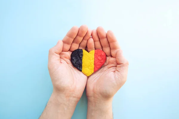 stock image Flag of Belgium. Heart shape of black, yellow and red plasticine modeling clay in male hands on blue background. Belgian flag concept, love. Top view, closeup, copy space.