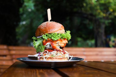 Uzun, lezzetli bir hamburger ya da sokak kafesinde, ahşap masada siyah tabakta sığır burger. Güneşli bir günde kahvaltı, fast food. Boşluğu kopyala, ön görünüm.