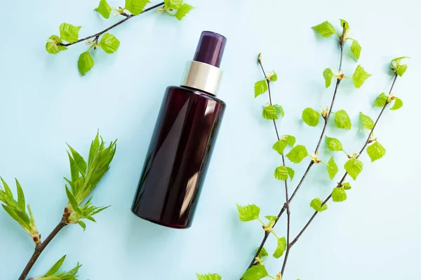 Brown cosmetic spray bottle and birch branches with young small leaves on blue background. Mockup. Skincare beauty and liquid antibacterial spray. Natural Body mist. Front view, flat lay.