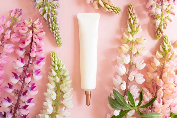 Face and eye cream squeeze cosmetic white tube with long nozzle and bronze screw cap and pink lupine flowers on pink background. Mockup, Cream, skincare, moisturizer, flat lay.