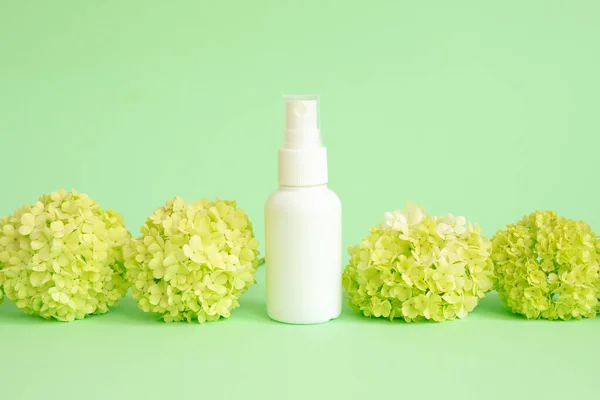 White cosmetic spray bottle and green viburnum flowers on green background. Front view, mockup, template. Natural organic spa cosmetics and liquid antibacterial spray concept.