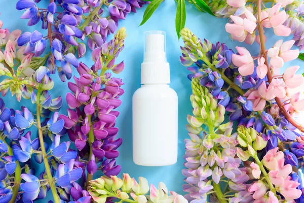 White cosmetic spray bottle and multicolored lupine flowers on blue background. Front top view, mockup, template. Natural organic spa cosmetics, liquid antibacterial spray, sanitizer, perfume