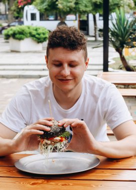 Kafkasyalı genç bir adam açık kafede siyah hamburger ya da sebze ve soslu sığır burger yiyor ve bakıyor. Güneşli bir günde kahvaltı, fast food..