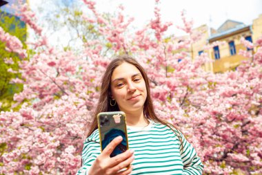 Kiraz çiçekli sakura arka planında akıllı telefondan selfie çeken kadın portresi. Genç beyaz kadın cep telefonuyla selfie portresi çekiyor. Yaz baharı vesikalık portresi.
