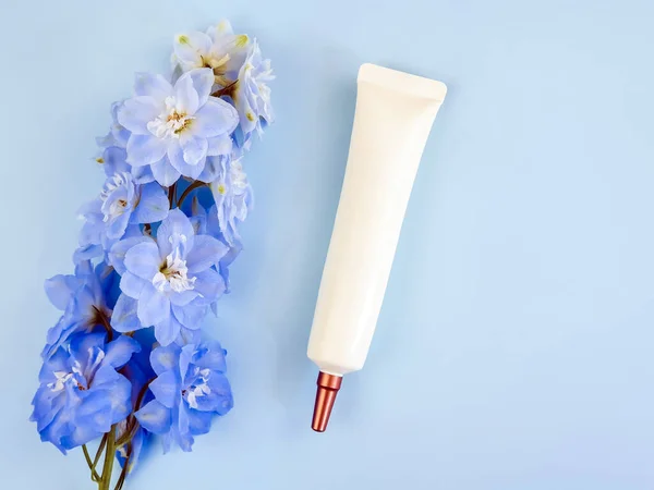 Face and eye cream squeeze cosmetic tube with long nozzle and bronze screw cap and blue Delphinium flower on blue background. Moisturizer blank bottle for branding - skincare, cream. Mockup