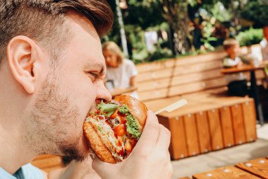 Genç, aç, beyaz bir adam açık kafede hamburger ya da sebzeli sığır burger ve sos yiyor. Güneşli bir günde brunch yapıyoruz. Yakın çekim, fast food, portre.