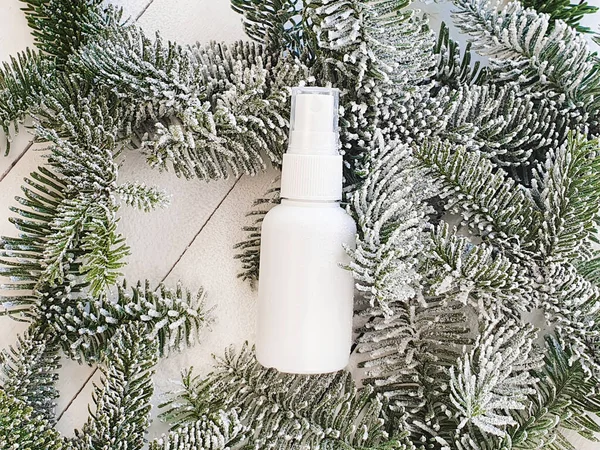 White cosmetic spray bottle and snowy branches of Christmas fir tree on white wood background. Mockup branding. Skincare beauty and liquid antibacterial spray. Natural Body mist. Front view, flat lay.